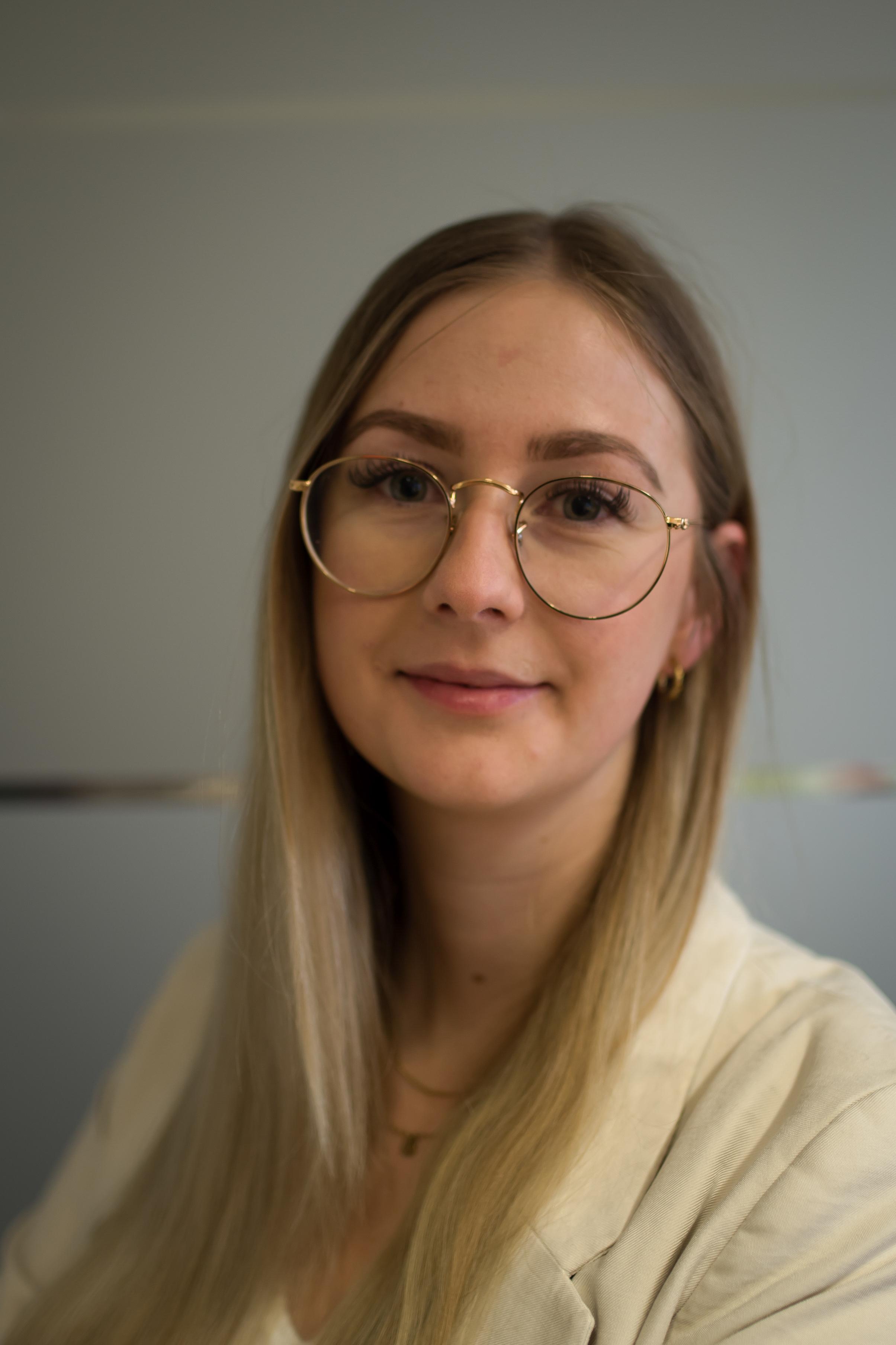 Junge Frau mit langen blonden Haaren und runder Brille lächelt freundlich